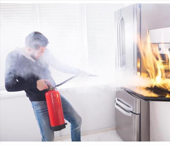 Young Man Using Red Fire Extinguisher To Stop Fire Coming From Oven In Kitchen.